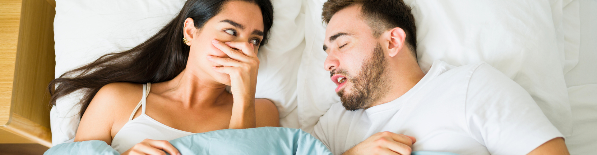 Woman in bed covering her nose facing man blowing bad breath.